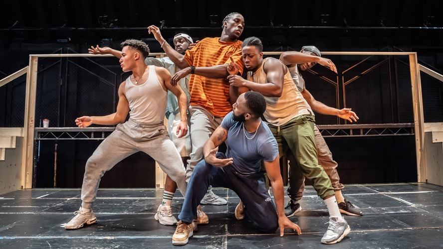 A group of performers rehearsing on stage, with scripts and musical instruments visible.