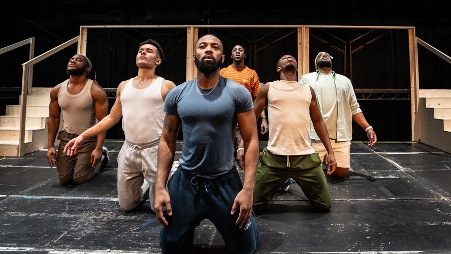A group of performers rehearses on stage, surrounded by props and stage lighting.