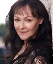 Frances Barber smiling, wearing a black dress, with shoulder-length hair and subtle makeup.