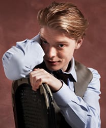 Freddie Fox smiling, wearing a dark suit and tie, with short, neatly styled hair.