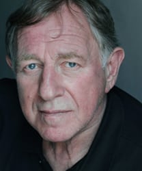 Geoffrey Freshwater smiling, wearing a dark suit and tie, against a neutral background.