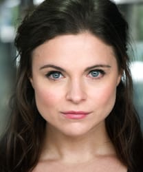 Gemma Sutton smiling, wearing a blue blouse, with shoulder-length brown hair and a neutral background.