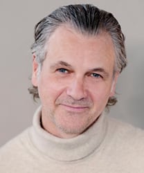 Guy Chambers smiling, wearing a black jacket, with short dark hair and a neutral background.
