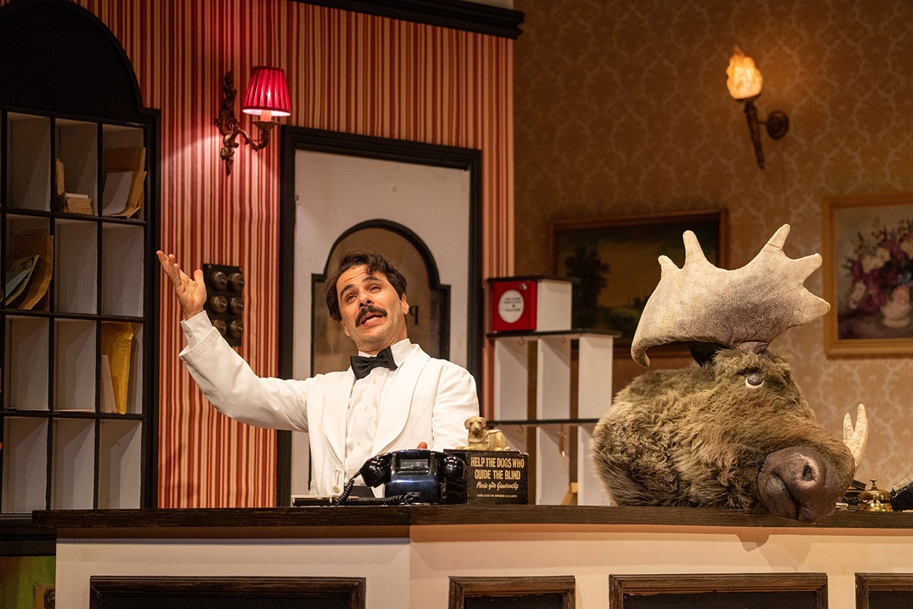 John Cleese as Basil Fawlty, wearing a suit, in a comedic pose on stage.