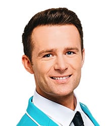 Harry Judd smiling, wearing a black shirt, with short brown hair and a neutral background.