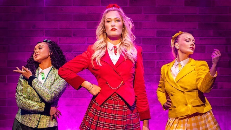 The cast of Heathers: The Musical poses together, dressed in vibrant costumes and smiling.