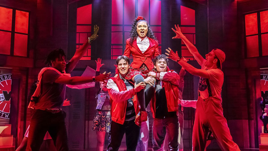 Cast of Heathers: The Musical performing on stage, dressed in vibrant costumes, with dramatic lighting.