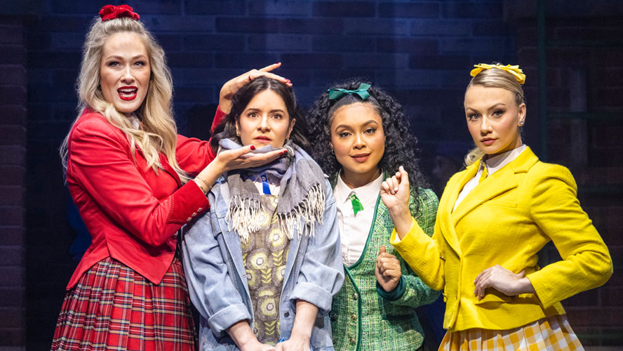 Cast members of Heathers: The Musical performing on stage, dressed in vibrant costumes with dramatic lighting.