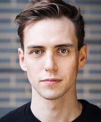 Jamie Muscato smiling, wearing a black shirt, with short dark hair and a neutral background.