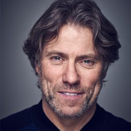 John Bishop performing on stage, wearing a black shirt, with a spotlight illuminating him.