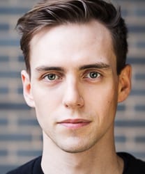 Jamie Muscato smiling, wearing a black jacket, with short dark hair and a neutral background.
