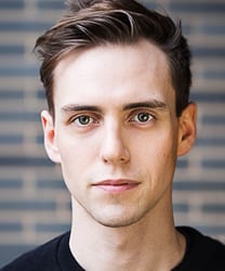 Jamie Muscato smiling, wearing a black shirt, with short dark hair and a neutral background.
