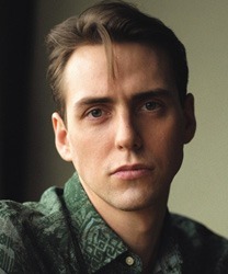 Jamie Muscato smiling, wearing a black shirt, with short dark hair and a neutral background.