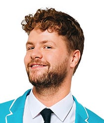 Jay McGuiness smiling, wearing a black jacket, with styled hair and a neutral background.