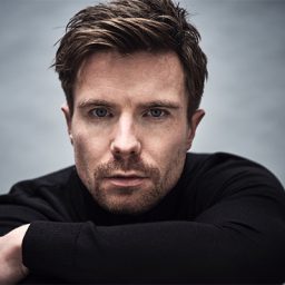 Joe Dempsie standing against a textured wall, wearing a casual black shirt and smiling.