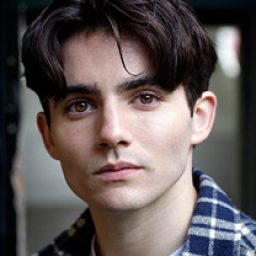 Jonny Amies smiling, wearing a casual shirt, with short brown hair and a neutral background.
