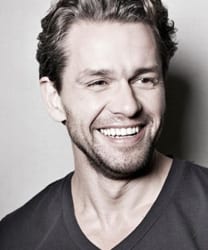 Julian Ovenden smiling, wearing a tailored suit, with short dark hair and a neutral background.