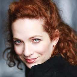 Katherine Parkinson standing on stage, smiling, wearing a black dress with a microphone in hand.