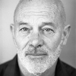 Keith Allen standing on stage, holding a script, with a spotlight illuminating him.