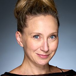 Katie Wimpenny smiling, wearing a black dress with a patterned overlay, standing against a neutral background.