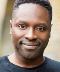 Kazeem Tosin Amore smiling, wearing a dark suit and tie, against a neutral background.