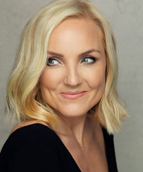 Kerry Ellis smiling, wearing a stylish black dress with elegant makeup and wavy hair.