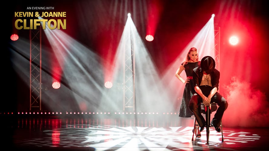 Kevin and Joanne Clifton smiling together, dressed in stylish outfits, against a vibrant stage backdrop.