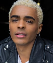 Layton Williams smiling, wearing a stylish outfit, with a blurred background of greenery.