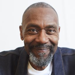 Lenny Henry performing on stage, wearing a black shirt, with a spotlight illuminating him.