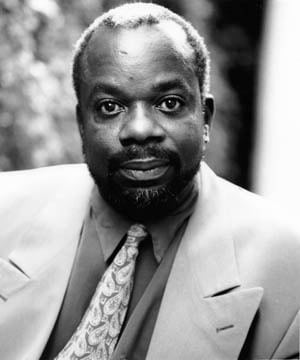 Joseph Marcell smiling, wearing a dark suit and tie, against a neutral background.