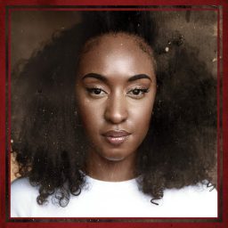 Lauran Rae with curly hair, wearing a white top, against a blurred brown background.