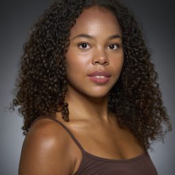 A young woman with curly hair wears a brown top, set against a dark gradient background.