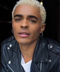 Layton Williams smiling, wearing a stylish outfit, with a vibrant background and soft lighting.
