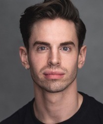 Liam Dean smiling, wearing a navy blue shirt, with short brown hair and a neutral background.