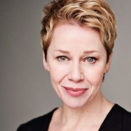 Linzi Hateley smiling, with shoulder-length hair and a black top against a neutral background.