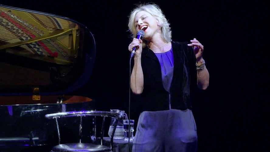 Liza Pulman smiling, wearing a stylish outfit, with a blurred background.