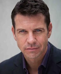 Lloyd Owen smiling, wearing a dark suit and tie, with a blurred background.