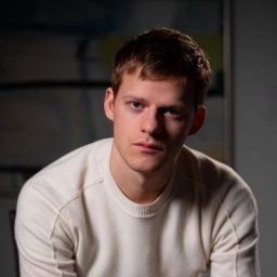 Lucas Hedges with short hair, wearing a black shirt, smiling against a neutral background.