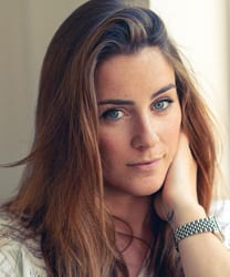 Lucie Jones smiling, wearing a stylish outfit, with long hair and a bright background.