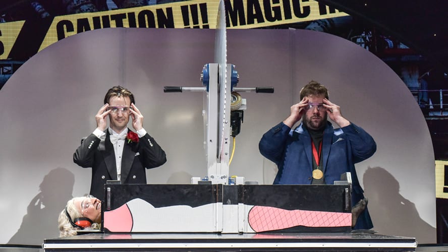 A stage scene featuring actors in vibrant costumes performing a comedic moment with props.