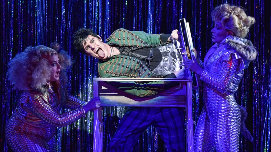 A stage scene featuring actors in vibrant costumes performing a comedic skit with props.