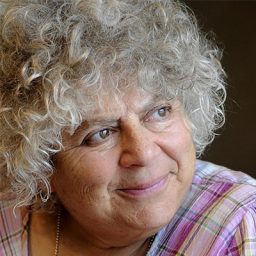Miriam Margolyes smiling, wearing a black top, with a blurred background of a stage.