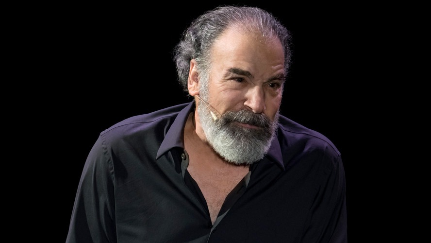 Mandy Patinkin smiling, wearing a dark suit and tie, with a blurred background.