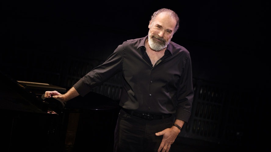 Mandy Patinkin smiling, wearing a dark suit and tie, against a blurred background.