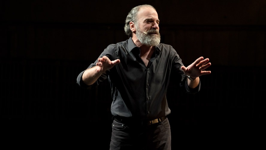 Mandy Patinkin smiling, wearing a dark suit and tie, with a blurred background.