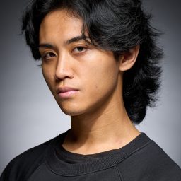 Young man with dark, wavy hair wearing a black sweatshirt, set against a grey background.