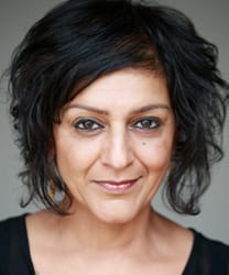 Meera Syal smiling, wearing a black top, with long dark hair and a neutral background.
