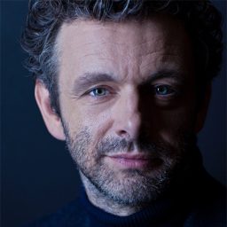 Michael Sheen smiling in a professional headshot, with a neutral background and soft lighting.