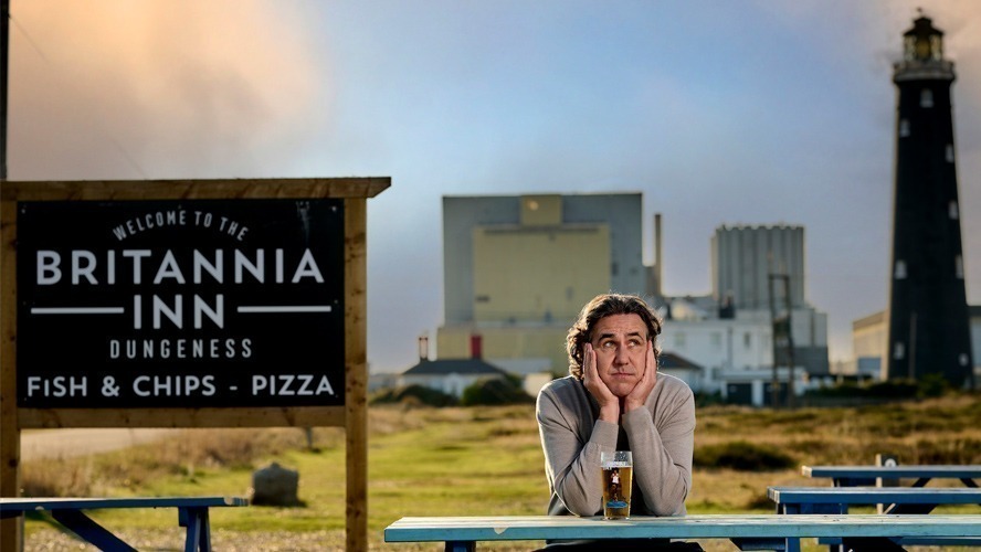 Micky Flanagan smiling, wearing a casual jacket, against a plain background.