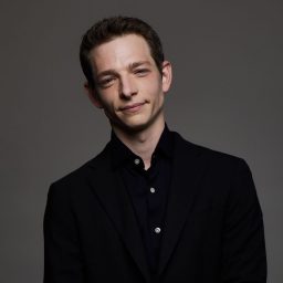 Mike Faist smiling, wearing a black shirt, with a blurred background.